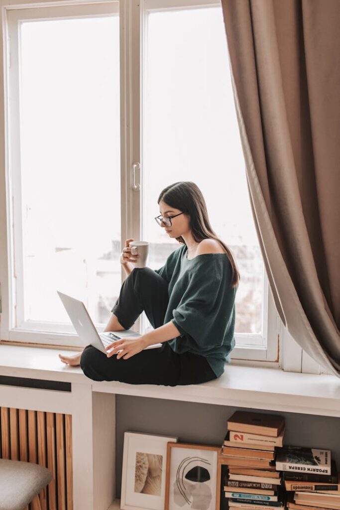 Side view full length of young concentrated female in dark casual clothes and eyeglasses sitting on windowsill with legs crosses and browsing netbook whlie enjoying cup of hot drink